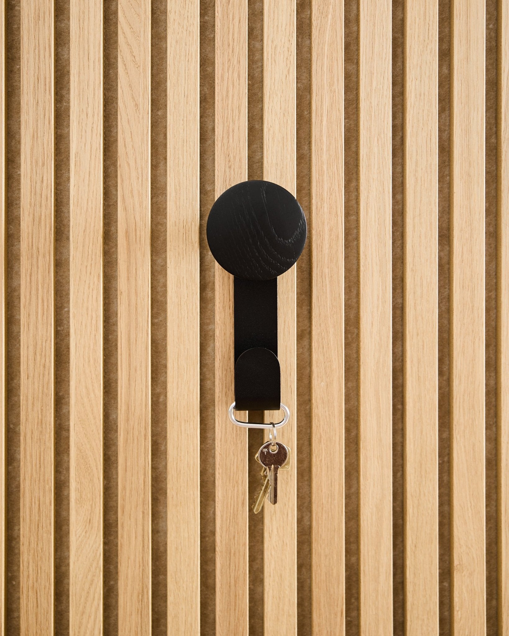 Un Linn Vertical Hanger en Black Oak de WoodUpp sostiene un llavero con dos llaves y una etiqueta redonda, montado sobre un panel acústico de madera Akupanel con textura.