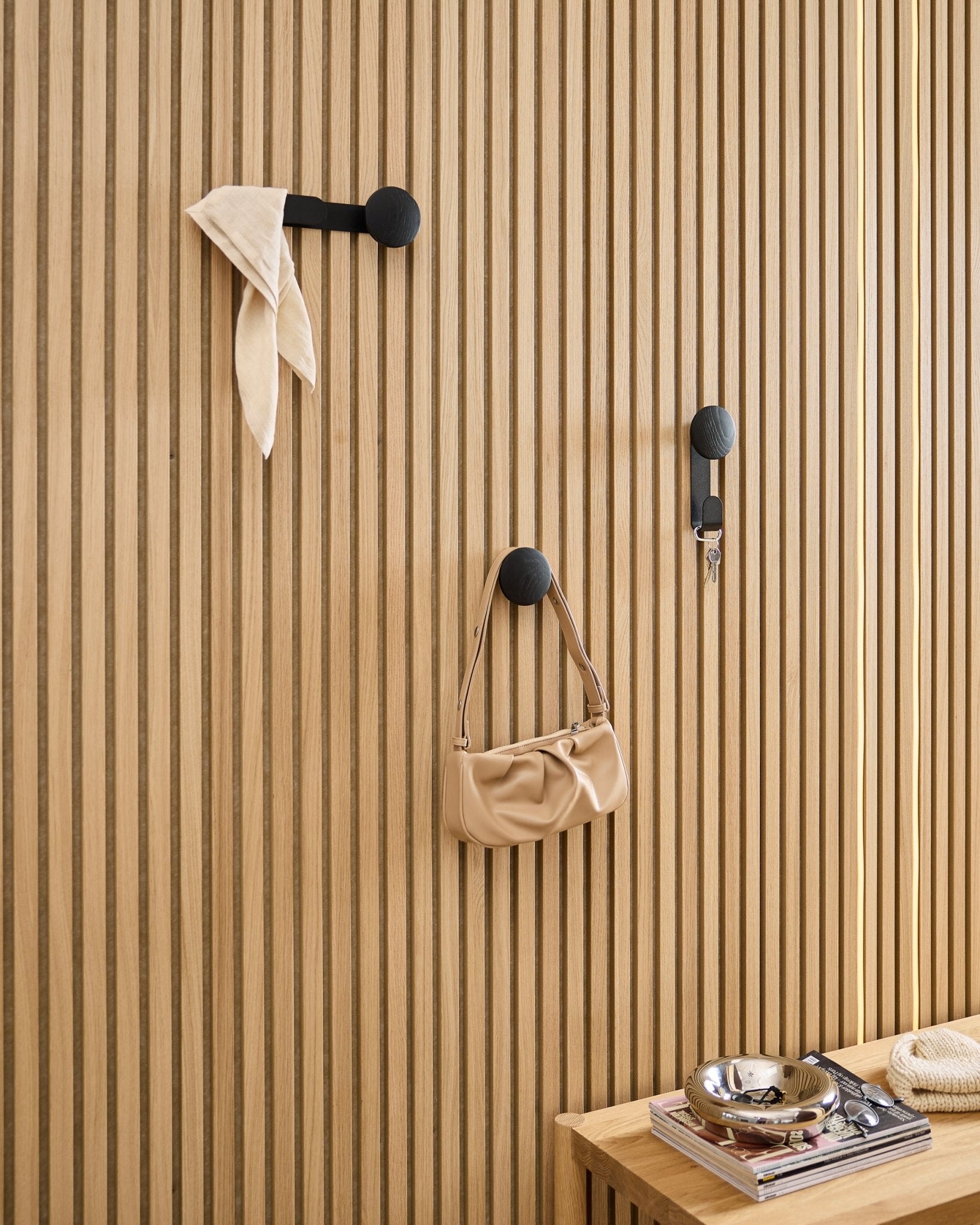 El Linn Single Hanger de WoodUpp en Black Oak sostiene una bufanda, un bolso beige y unas llaves sobre un panel acústico de madera de lamas black Akupanel; cerca hay un banco con libros y una bandeja metálica.