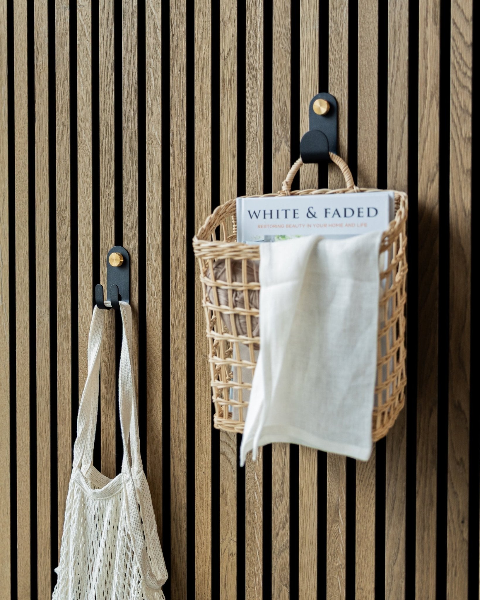 El Norr Single Hanger de WoodUpp en Black, con una cesta tejida y una bolsa de malla, está montado en un panel acústico nórdico de listones para mantener el orden.
