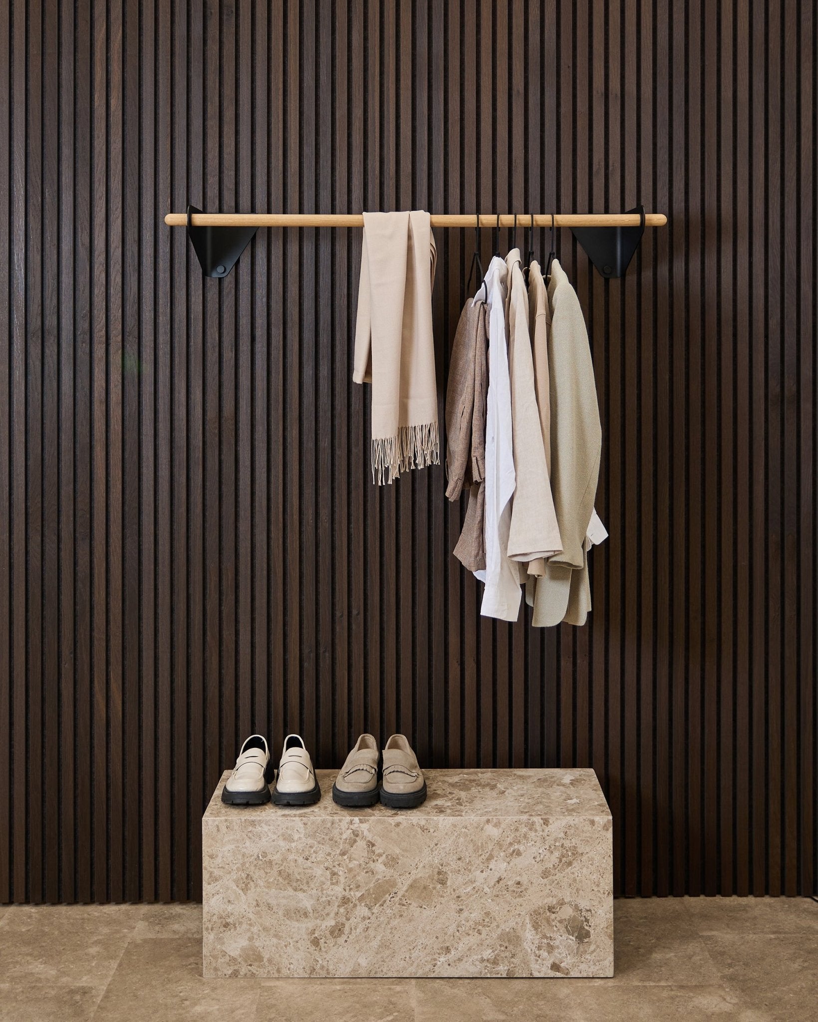Prendas en tonos neutros y una bufanda cuelgan de un black de 115,9 cm Linn | Coat Rail de WoodUpp sobre un banco de mármol, frente a un panel acústico de lamas Akupanel oscuro.