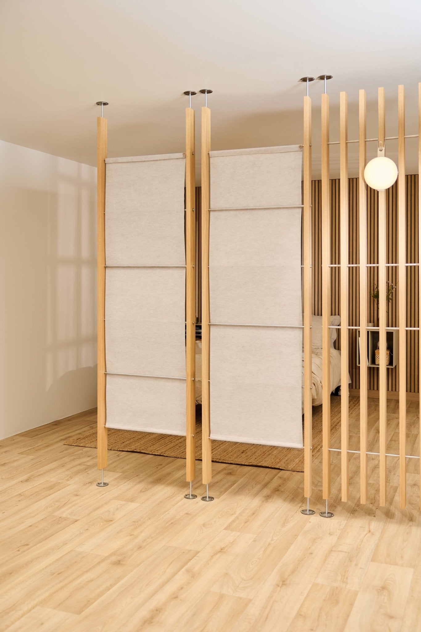 Habitación moderna con suelos de madera clara, un separador de ambientes de fieltro WoodUpp EMBRACE en color beige claro, una cama y una lámpara de pared redonda visible al fondo.