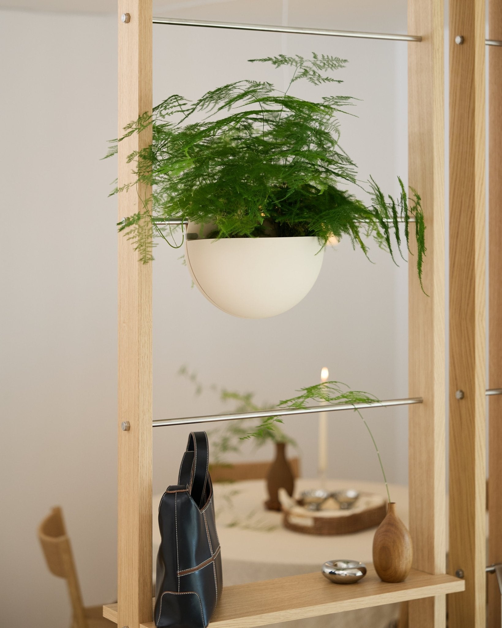 Un helecho verde, colocado en una maceta WoodUpp Gro Soft Pocket BM2 de color beige claro, descansa sobre una estantería de madera, encima de una bolsa negra, un jarrón y una mesa de comedor puesta.