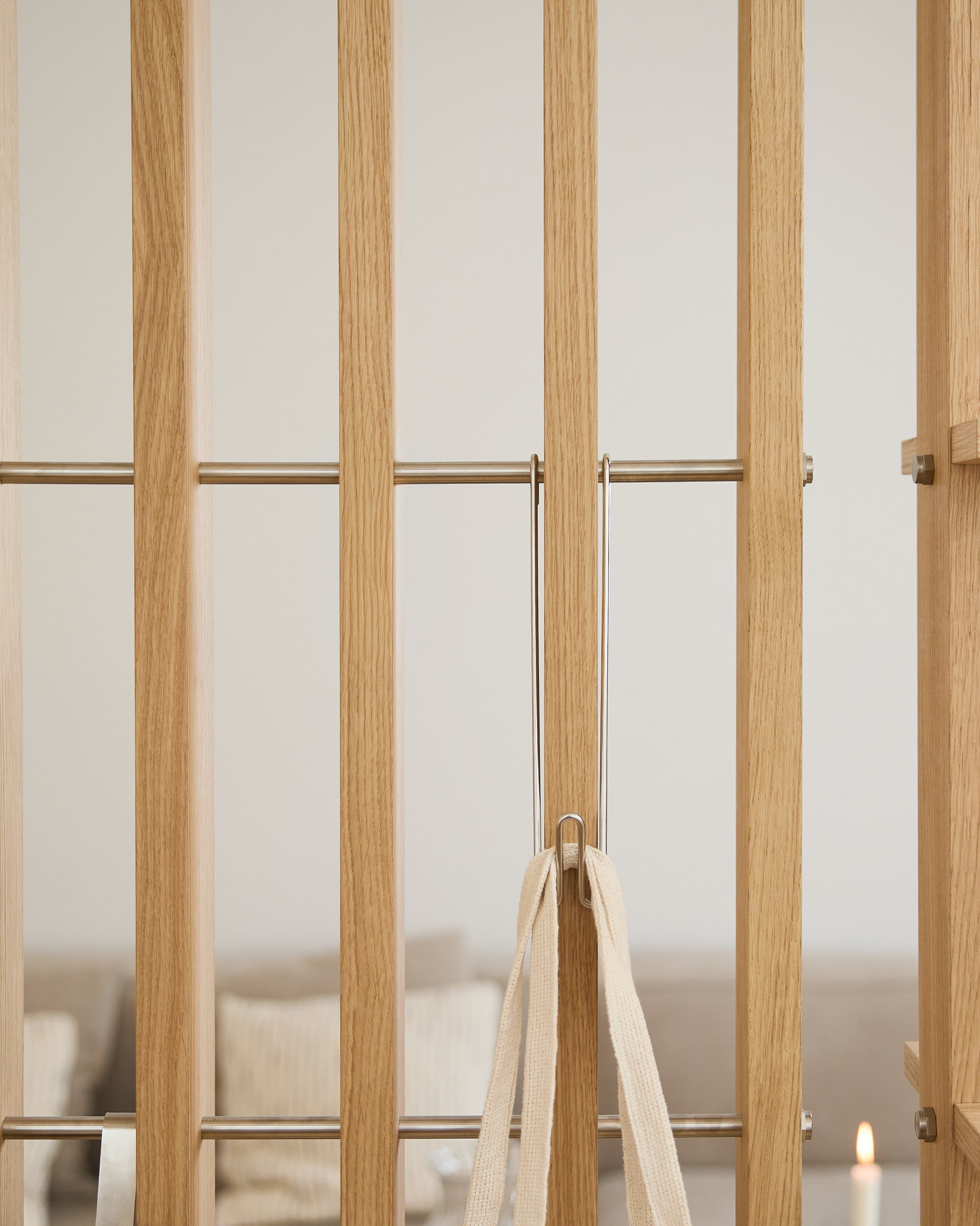 Puerta de madera clara con barras metálicas, un lazo beige y tres ganchos WoodUpp Alva Wire Hooks/BM1; sofá y vela difuminados al fondo.