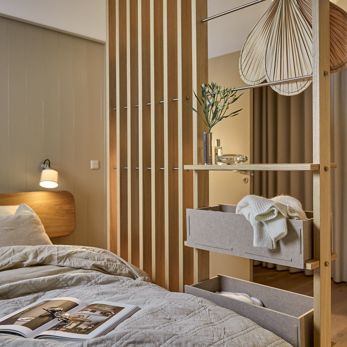 Un dormitorio moderno con ropa de cama nórdica suave, lámpara colgante y una revista sobre la cama cuidadosamente hecha. Los paneles de madera acústica WoodUpp Akupanel acentúan la pared.