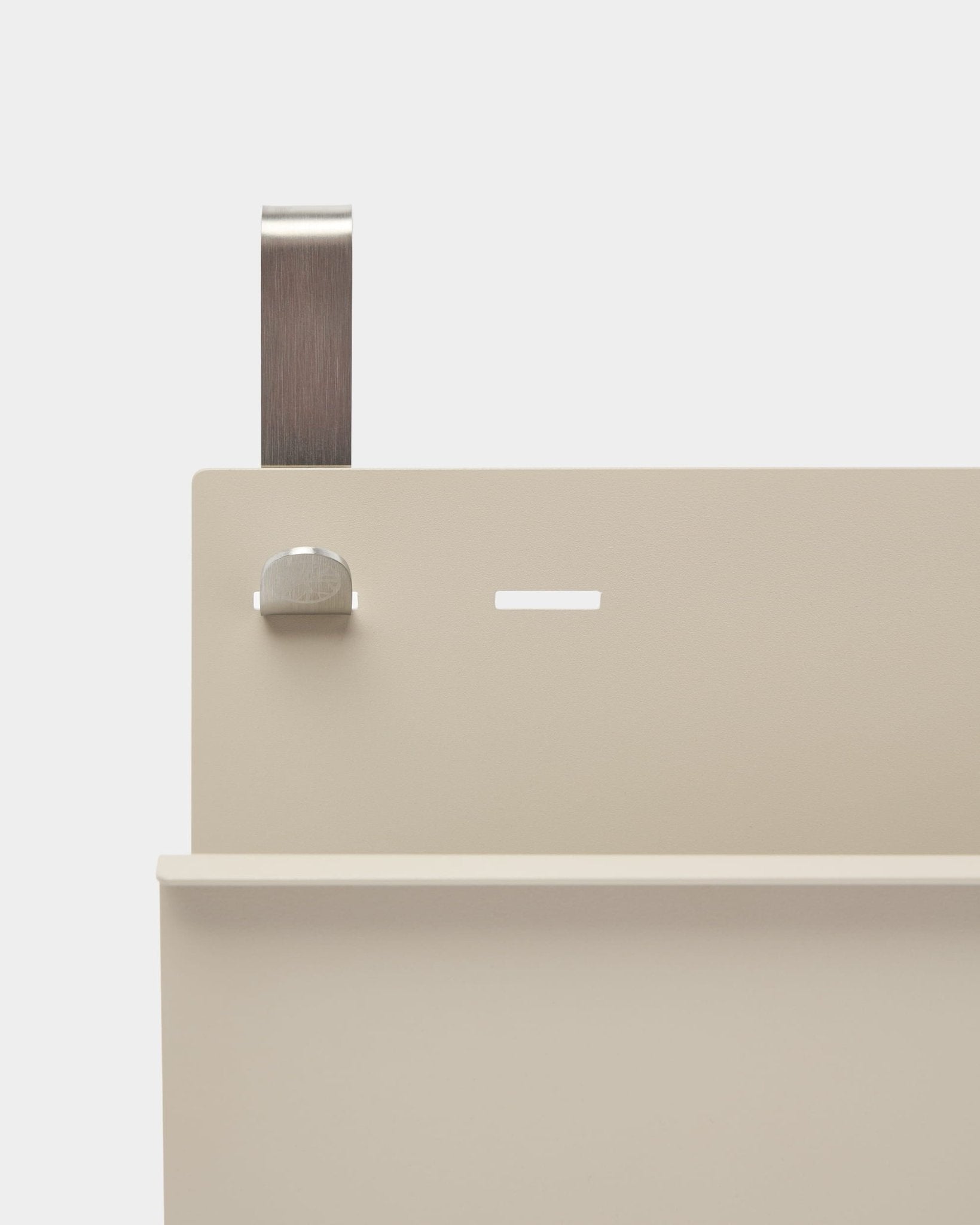 Primer plano del revistero Nell de WoodUpp en beige, de steel con recubrimiento en polvo, con balda, ranura, tornillo y asa curva; funciona como separador de ambiente o revistero.