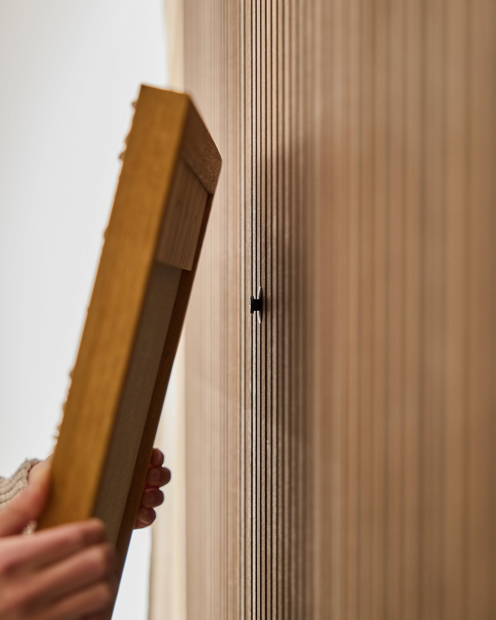 Una persona utiliza el set Dupps | Original de WoodUpp para montar un marco de panel acústico de madera Akupanel, centrándose en fijar un gancho small black cerca de las lamas vertical.