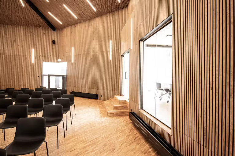 Amplia sala de conferencias con paneles acústicos de madera Akupanel en paredes y techo, hileras de sillas negras, grandes ventanales y luminarias verticales.