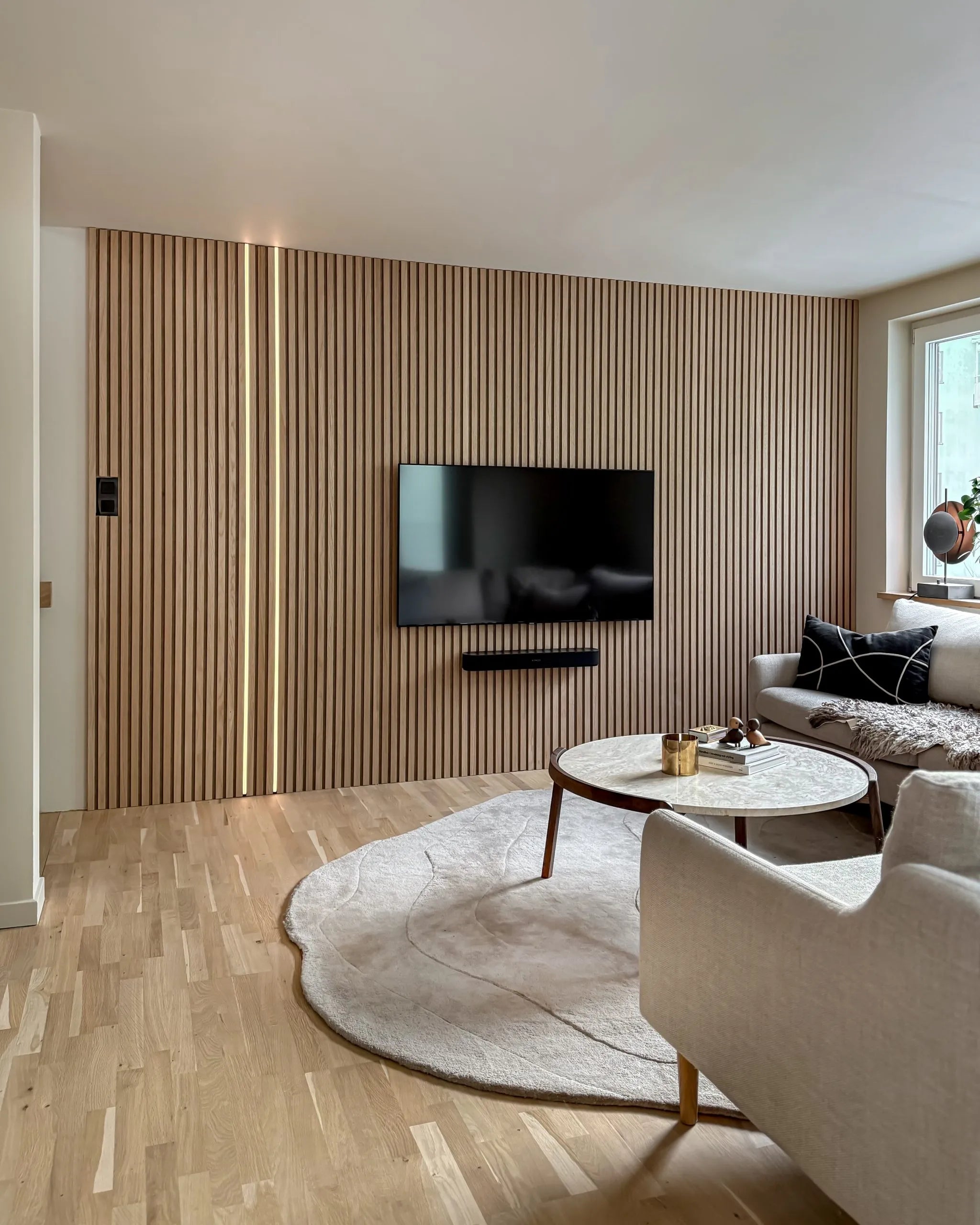 Moderno salón con una pared de acento nórdico de lamas de madera acústica Akupanel, TV montada, mesa de centro redonda, sofá crema, alfombra grande sobre suelos de madera clara.