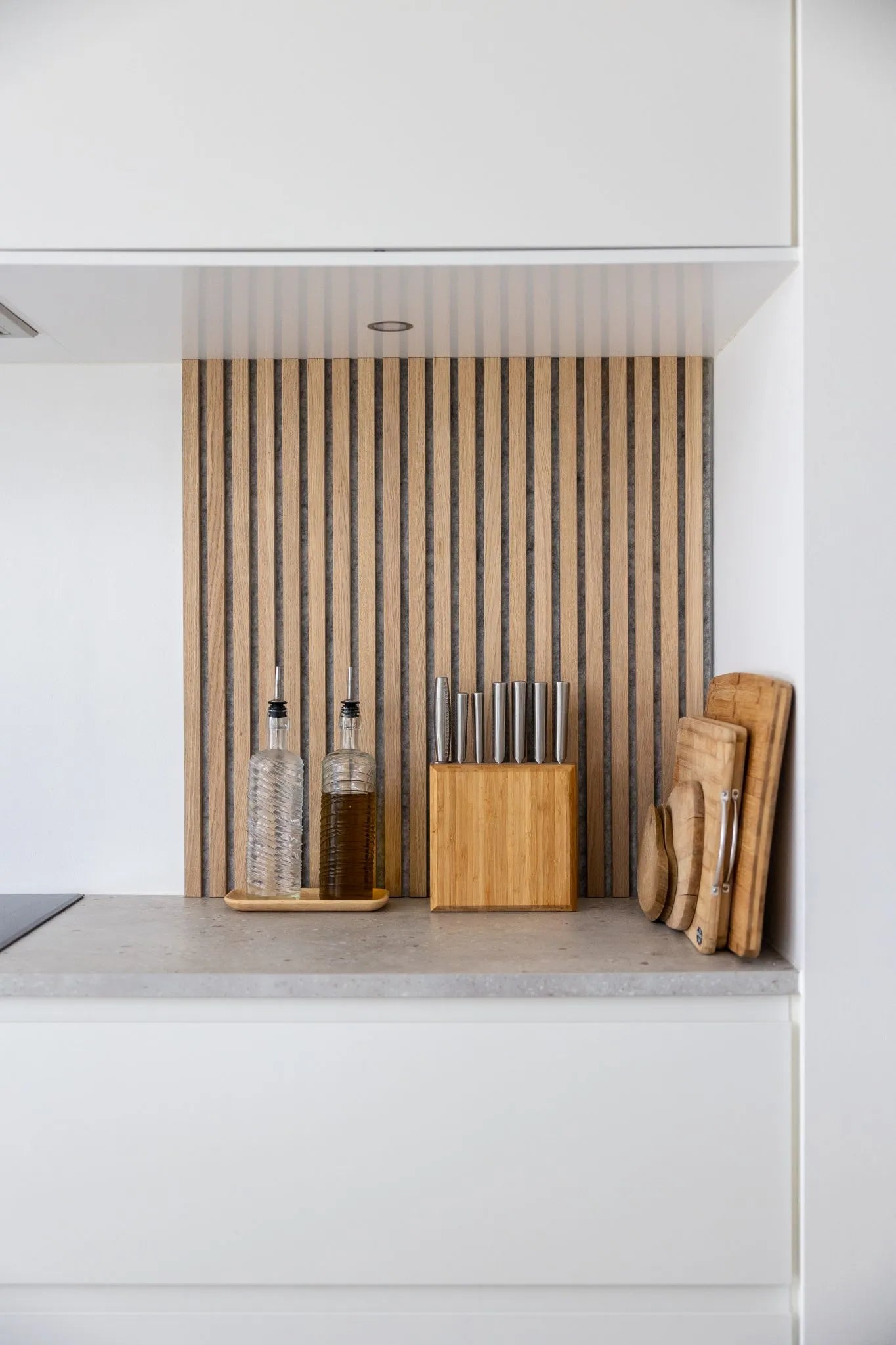 Encimera de cocina moderna con botellas de aceite, bloque de cuchillos de madera y tablas de cortar apiladas sobre un panel trasero de lamas de madera acústica Akupanel nórdica y armarios blancos.