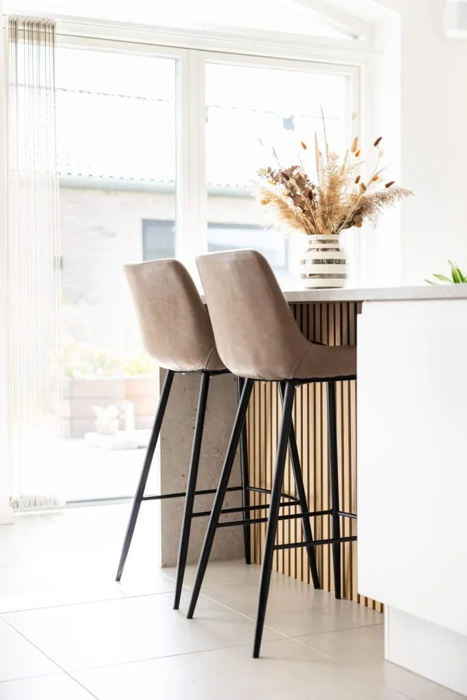 Taburetes de bar beige en una isla de cocina con un jarrón de flores secas, grandes ventanales y estilo nórdico contemporáneo con paneles acústicos en las paredes.