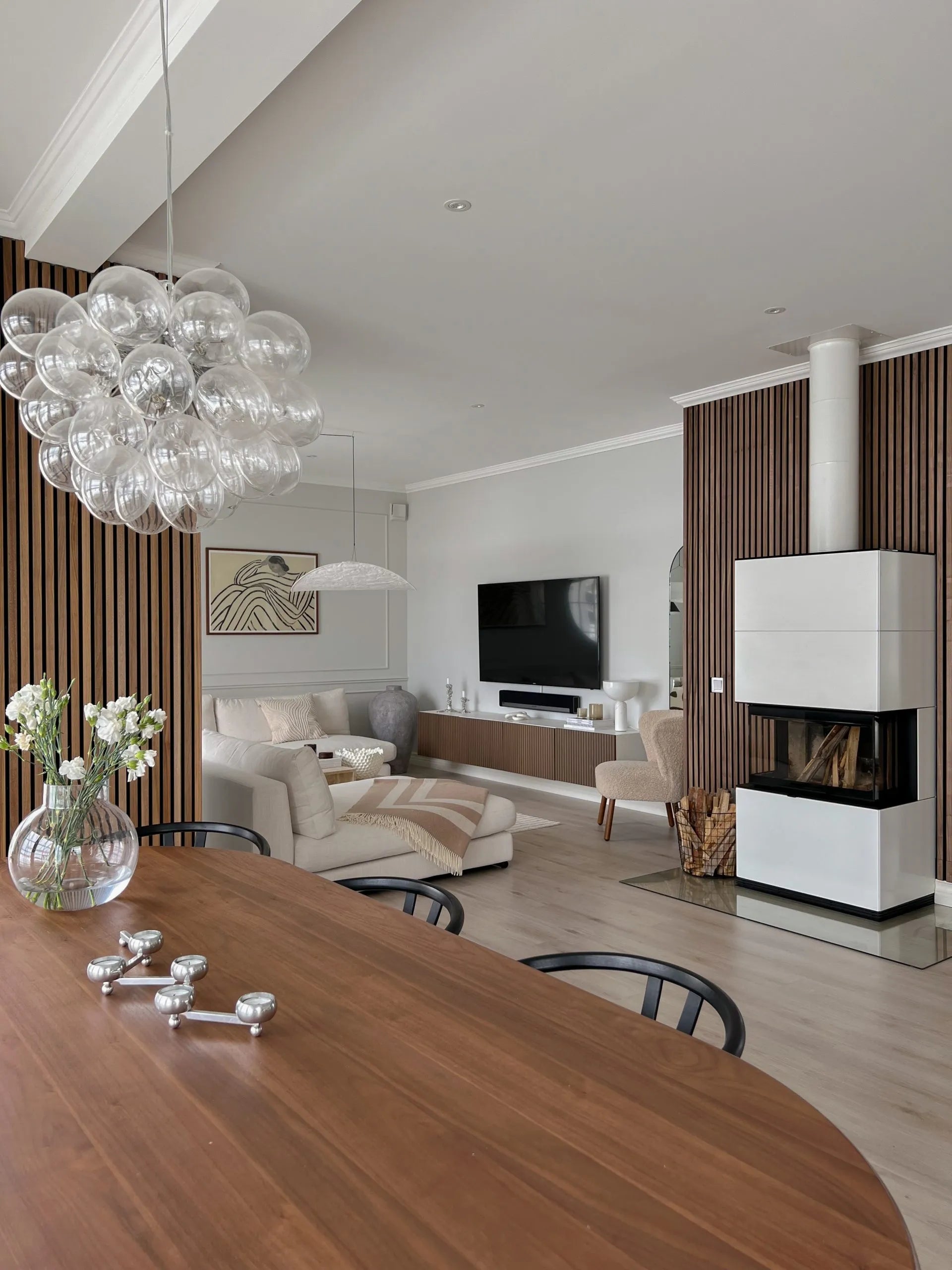 Moderna zona de estar y comedor con paneles acústicos de madera nórdica, acogedores asientos, TV, elegante chimenea con leña apilada y una araña de burbujas de cristal.
