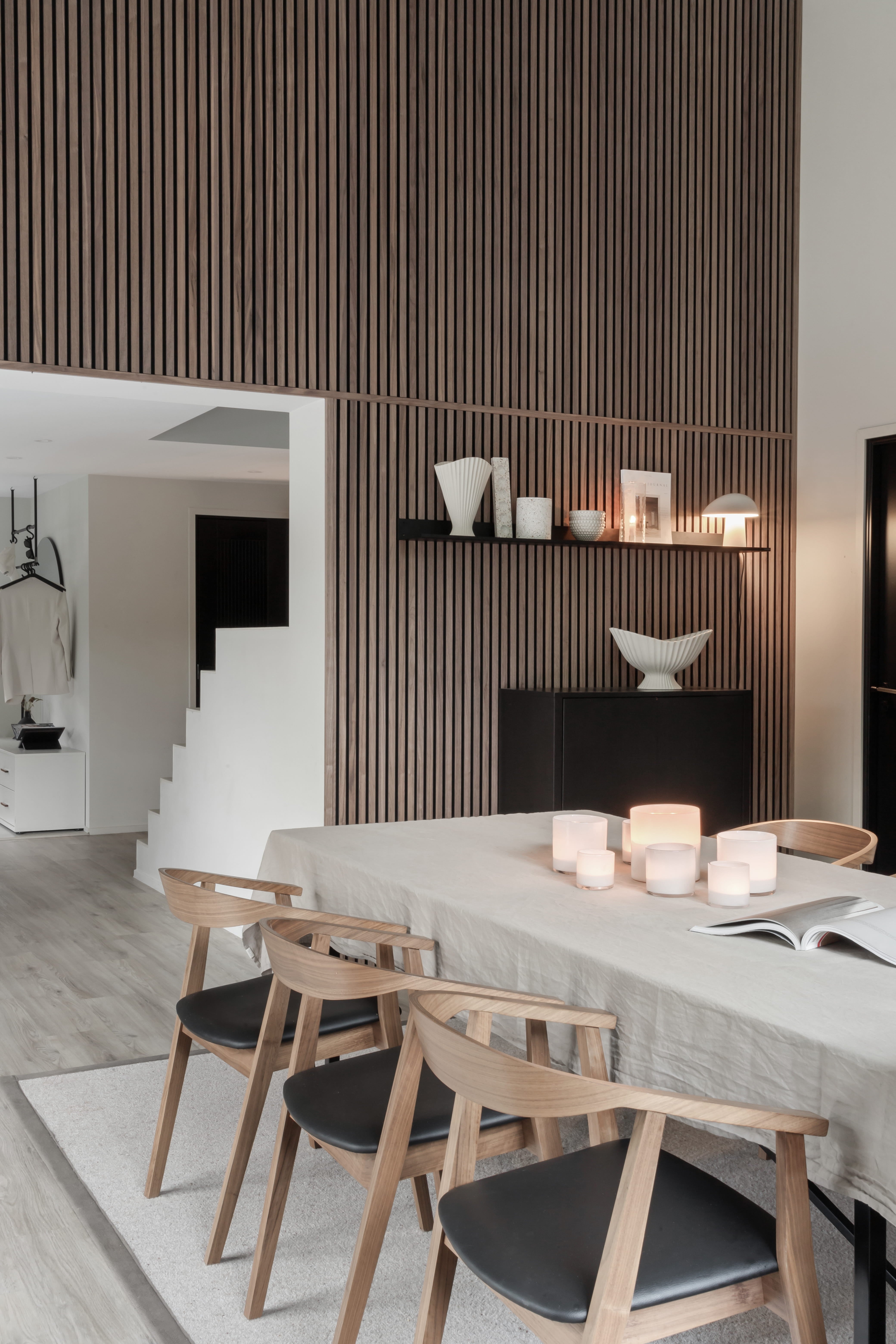 Comedor moderno con mesa de madera clara, cuatro sillas, velas y decoración minimalista. Pared vertical de lamas de madera acústica Akupanel y escalera abierta a la vista.