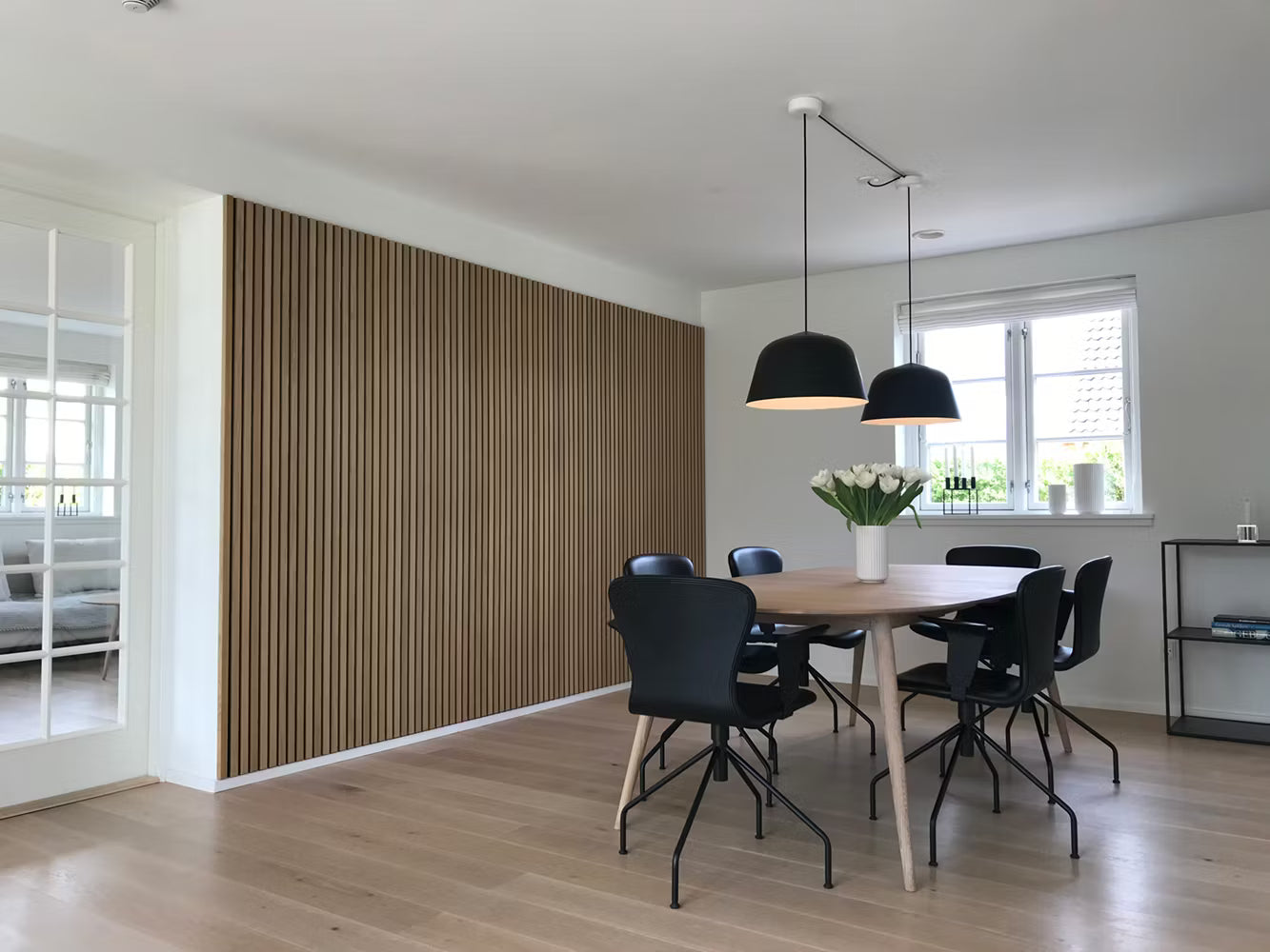 Comedor moderno con suelo de madera, acento de lamas acústicas de Akupanel, sillas negras, mesa de madera, lámparas colgantes y un centro de mesa floral blanco.