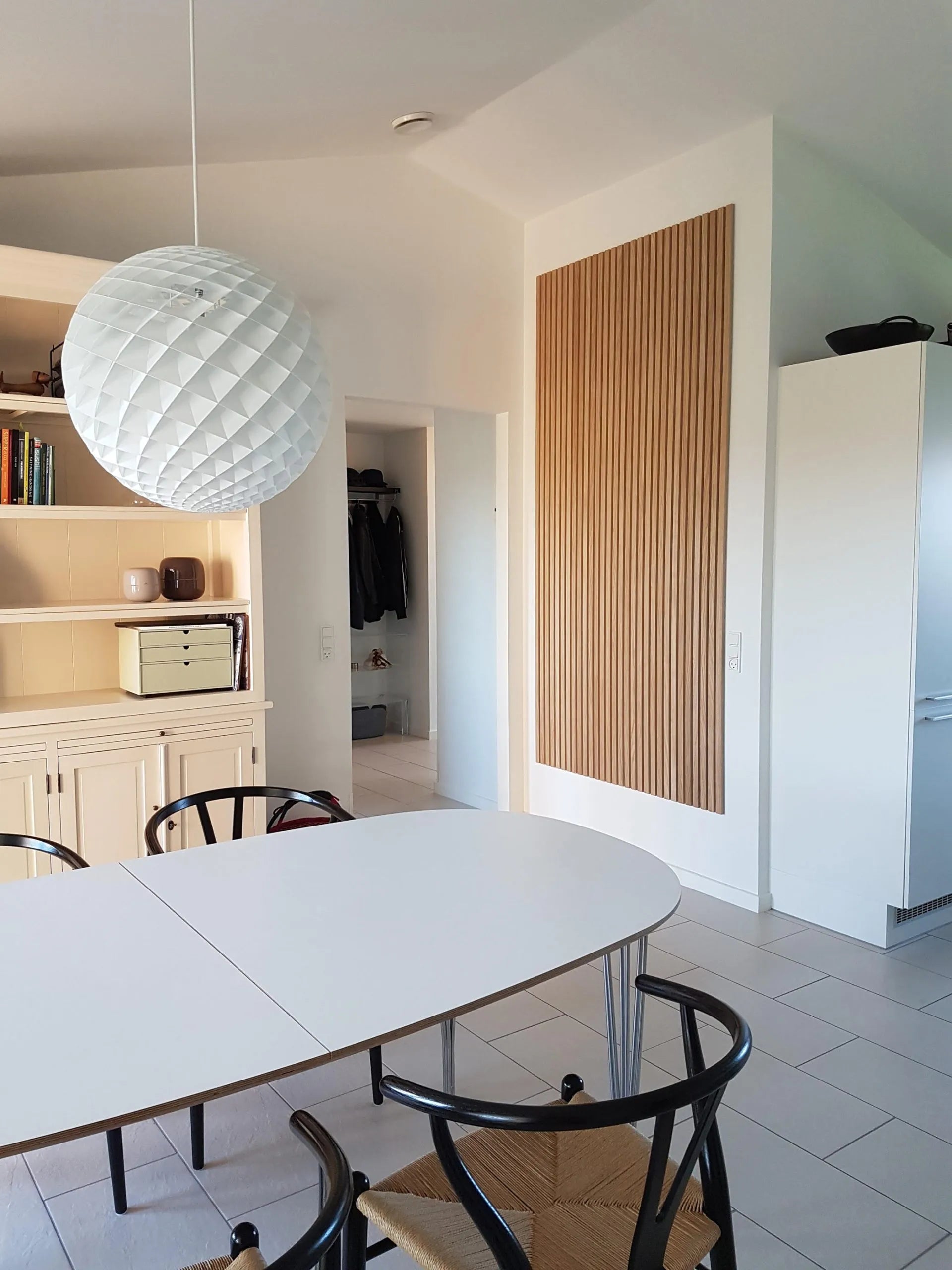 Comedor moderno con mesa ovalada blanca, sillas negras, gran lámpara colgante con textura y elegantes armarios acústicos de Akupanel de madera clara.