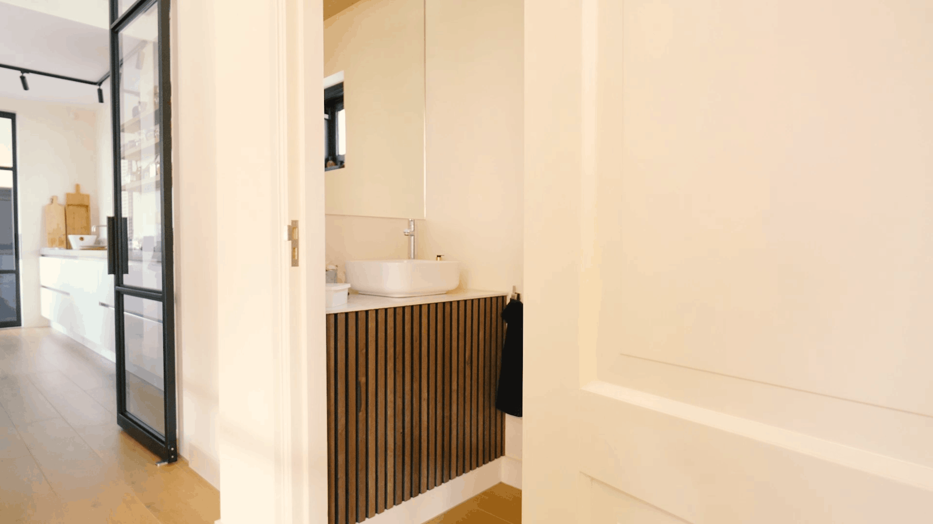 Cuarto de baño moderno con lavabo redondo, tocador acústico de listones de madera, espejo grande y puerta blanca junto a un pasillo luminoso y una cocina contemporánea.