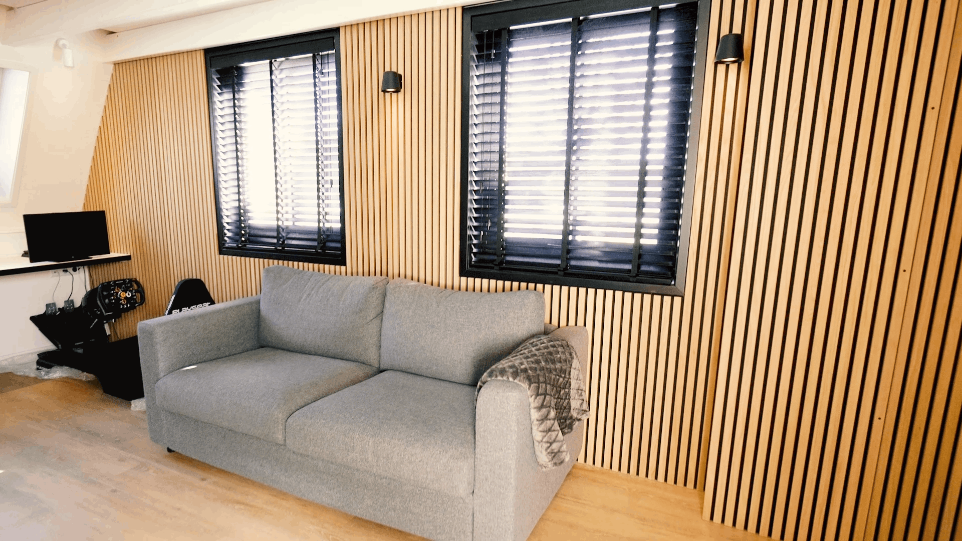 Sofá gris con manta estampada colocado frente a una pared de lamas de madera acústica Akupanel nórdica, flanqueada por dos ventanas con persianas negras y un escritorio compacto.