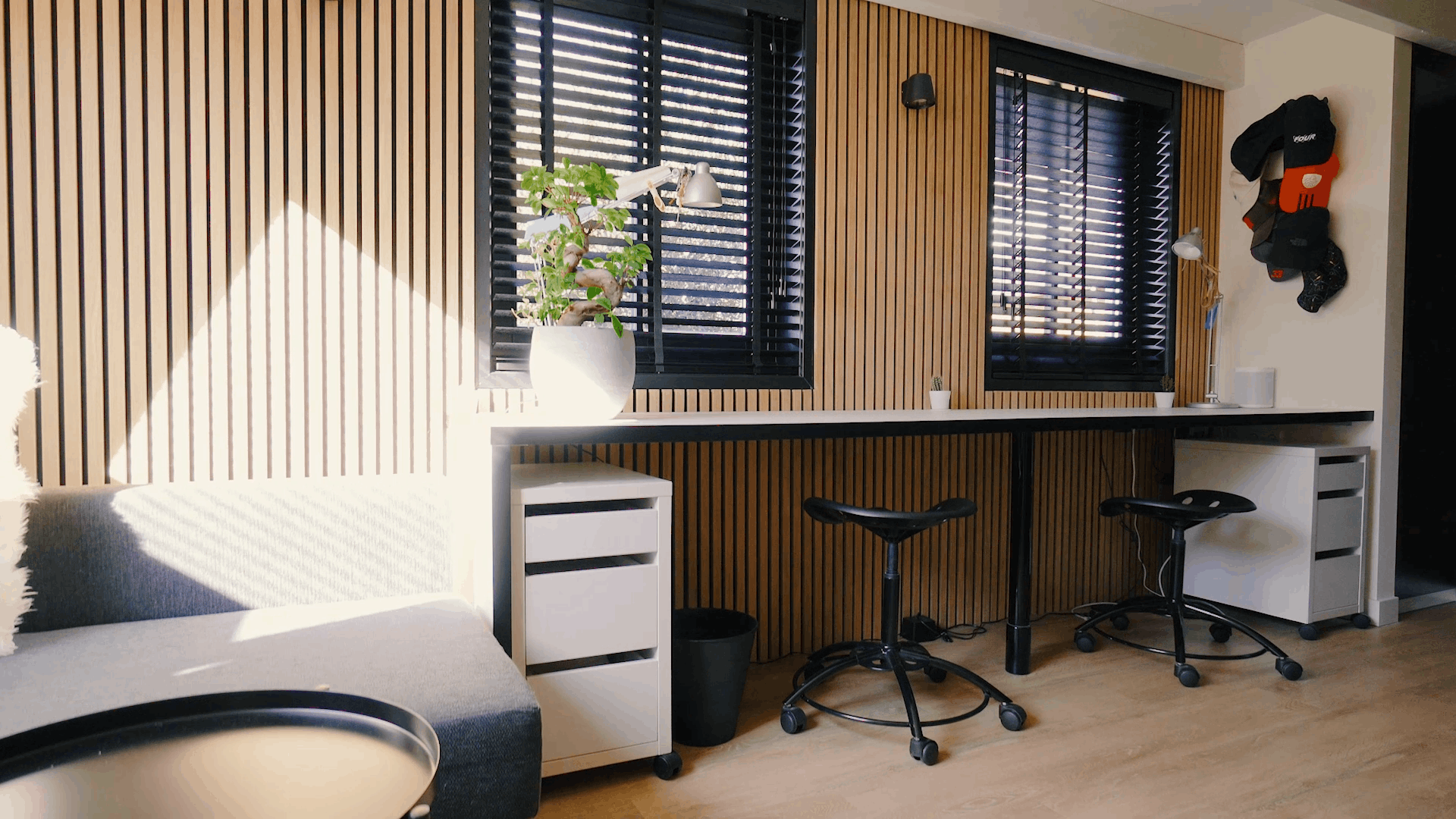Oficina moderna con paneles de madera acústica nórdica en las paredes, puestos de trabajo ergonómicos, taburetes negros, una planta en maceta y luz solar a través de las persianas negras de las ventanas.