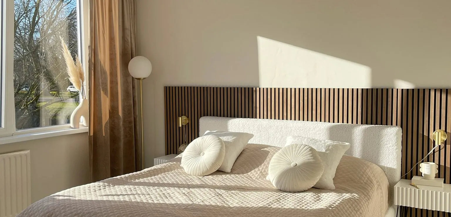 Dormitorio moderno iluminado por el sol con cama acolchada beige, almohadas redondas, cabecero de lamas de madera de Akupanel y decoración minimalista junto a una gran ventana con cortinas transparentes.
