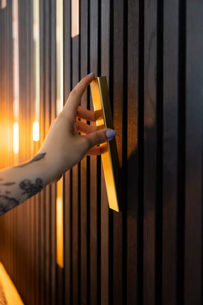 Una mano tatuada con uñas de color púrpura claro agarra un tirador dorado en una elegante pared oscura de paneles de madera acústica Akupanel, resaltada por una cálida iluminación.