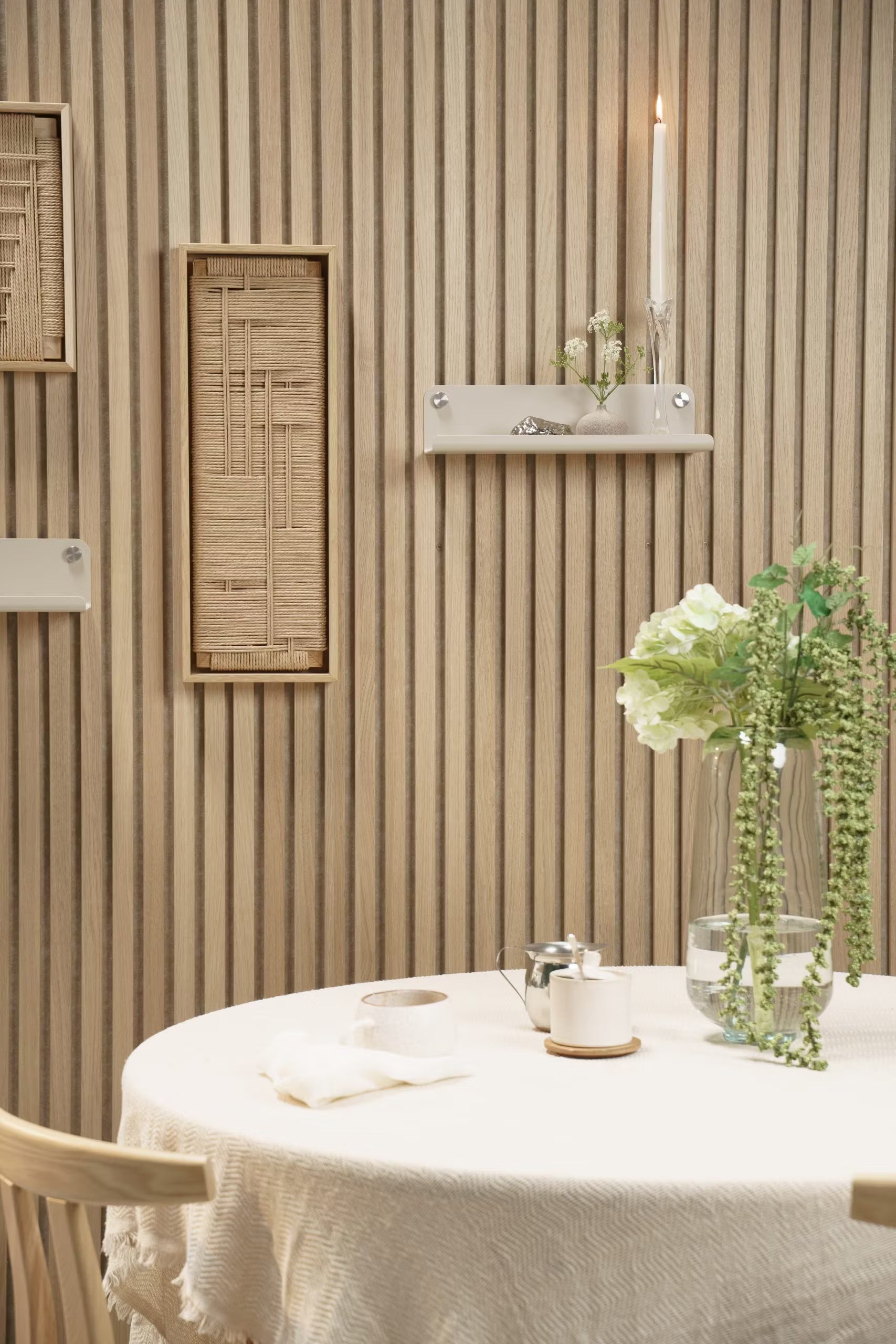 Una mesa redonda con un mantel blanco, un jarrón de flores, una vela y un adorno colocados delante de un tabique acústico de Akupanel de madera clara con detalles de tejidos y estantes.