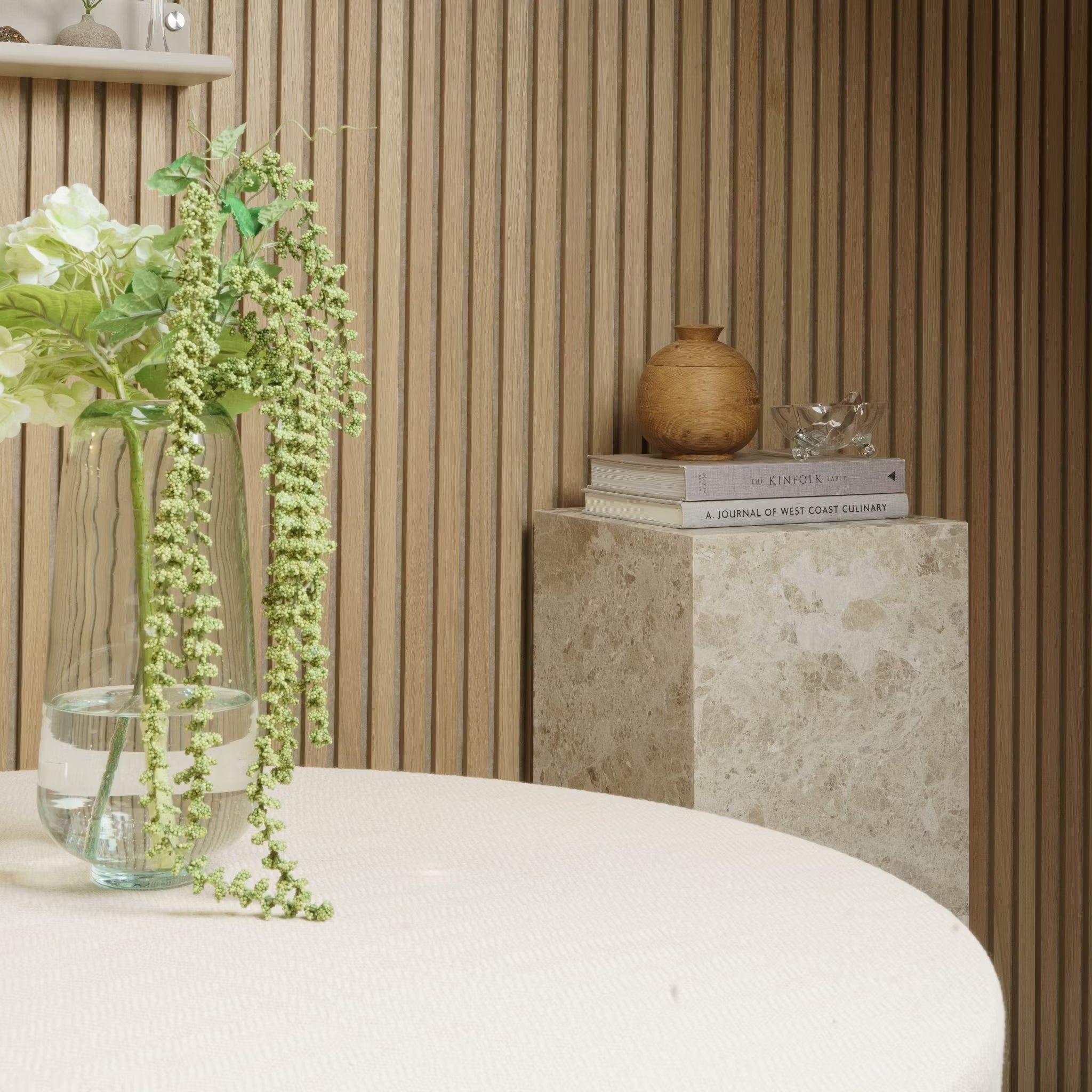 Un jarrón con flores descansa sobre una mesa redonda; libros y cerámica reposan sobre un pedestal de mármol contra una pared de paneles de madera acústica Akupanel.