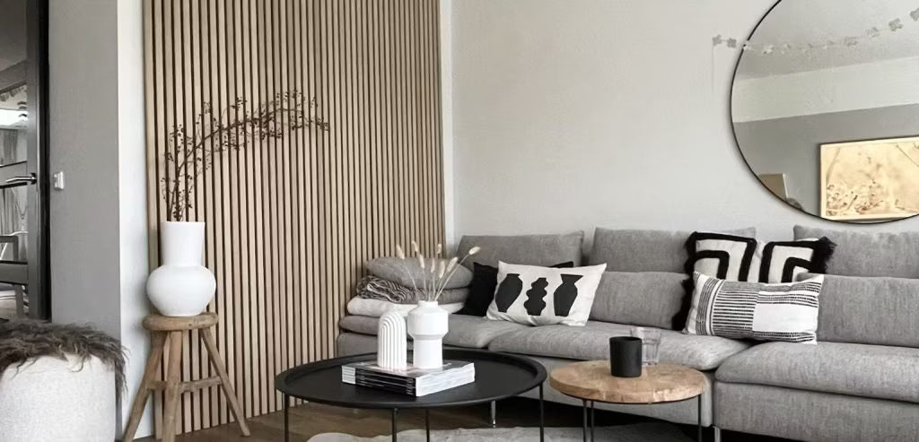 Sala de estar moderna con un sofá gris, cojines abstractos, mesas de centro negras, jarrones decorativos y un panel de lamas acústicas de madera de Akupanel con un espejo redondo.
