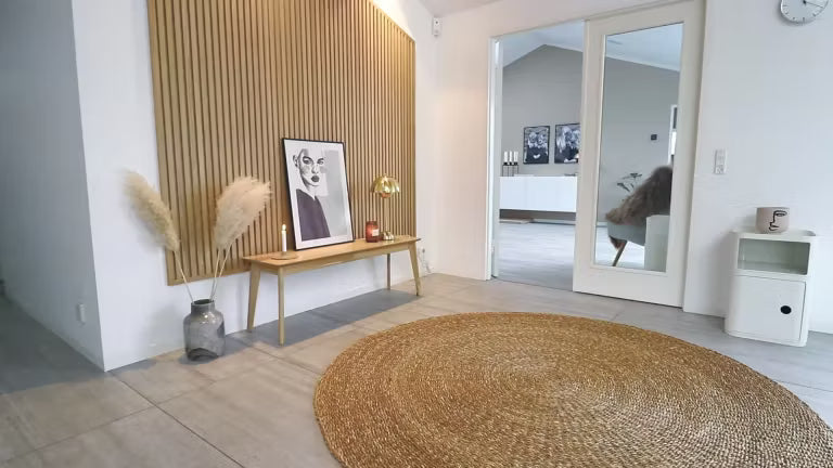 Salón nórdico minimalista con una alfombra redonda de yute, un banco de madera con hierba de la pampa, obras de arte enmarcadas y una pared acústica de listones de madera de Akupanel.