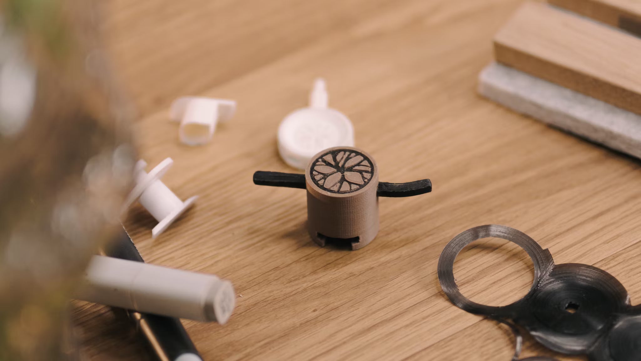 Un pequeño prototipo impreso en 3D con brazos negros descansa sobre un escritorio de madera junto a varios componentes de plástico, en referencia al desarrollo de accesorios para paneles acústicos.