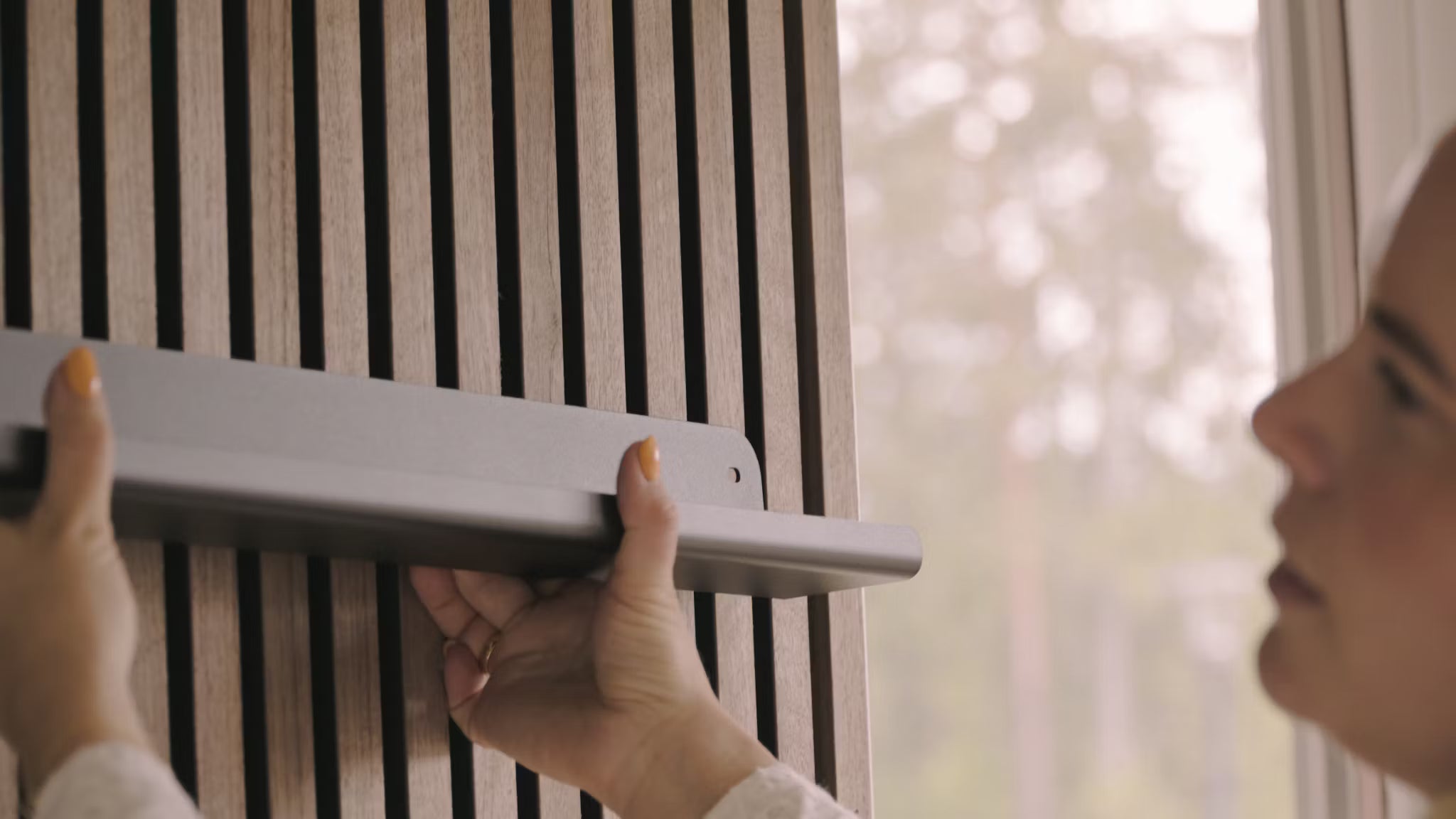 Una persona instala una estantería gris en una pared de lamas de madera acústica Nordic Akupanel junto a una ventana con vistas a los árboles.