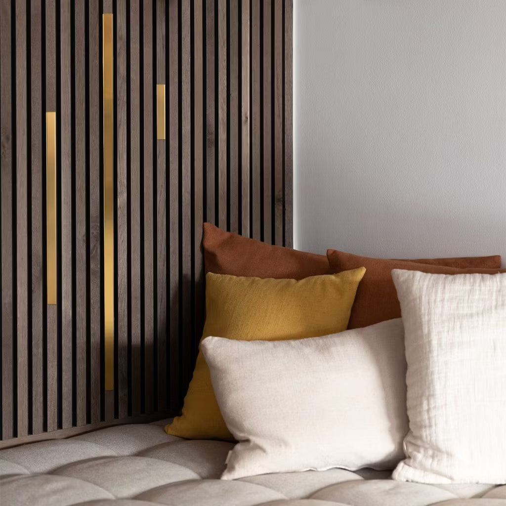 Cojines de color crema, amarillo y marrón sobre un sofá de pelo insertado beige junto a un moderno panel acústico de lamas con detalles dorados para un ambiente acogedor y contemporáneo.