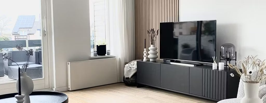 Moderno salón con mueble de TV negro, TV de pantalla plana, jarrones decorativos y grandes ventanales con luz natural que dan al patio; Akupanel en una pared.