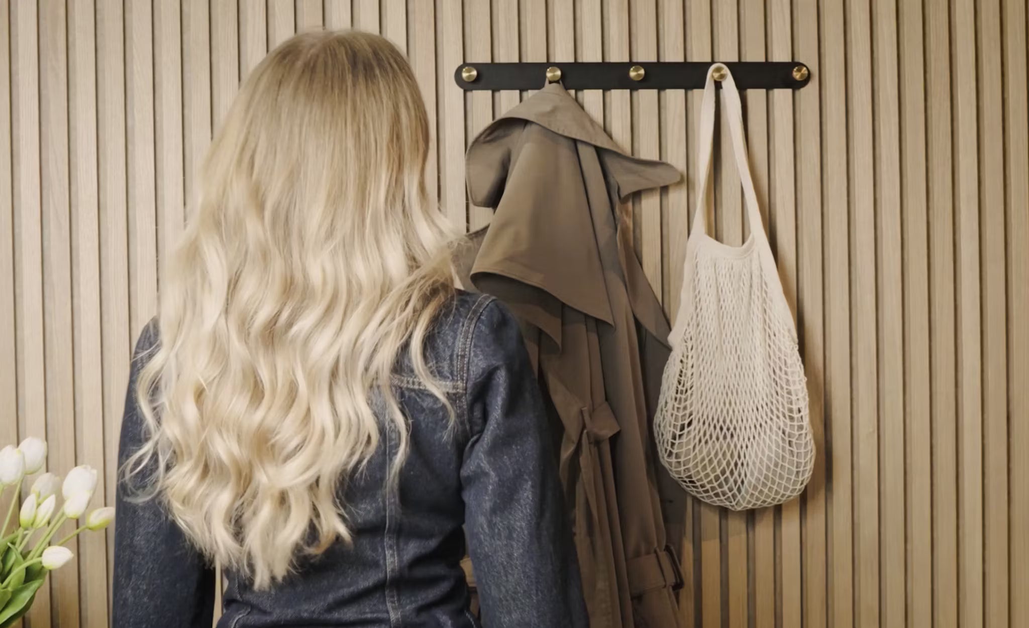 Una mujer de pelo largo y rubio está de pie frente a un perchero con una gabardina, una bolsa de red blanca y tulipanes blancos frescos junto a un panel de madera acústica Akupanel en la esquina.