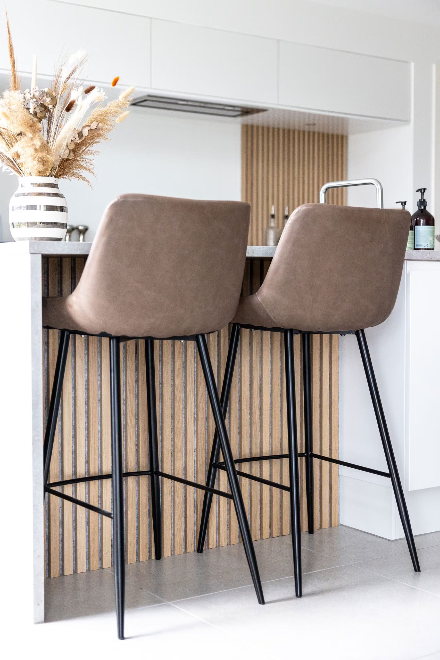 Taburetes de bar marrones con patas negras en una isla de cocina, con un panel de listones de madera acústica nórdica y un jarrón con flores secas sobre la encimera.