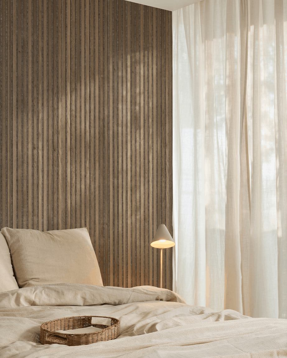 Dormitorio minimalista con ropa de cama beige, una bandeja de mimbre, un panel acústico de listones WoodUpp Akupanel Rustic Grey Oak, una lámpara blanca y cortinas translúcidas.
