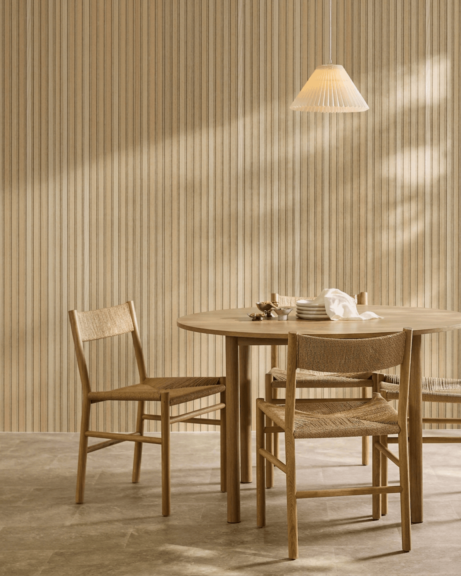 Comedor minimalista con una mesa redonda, sillas de mimbre y una lámpara colgante junto a un panel acústico de madera WoodUpp Akupanel en Roble Clásico con fieltro beige.