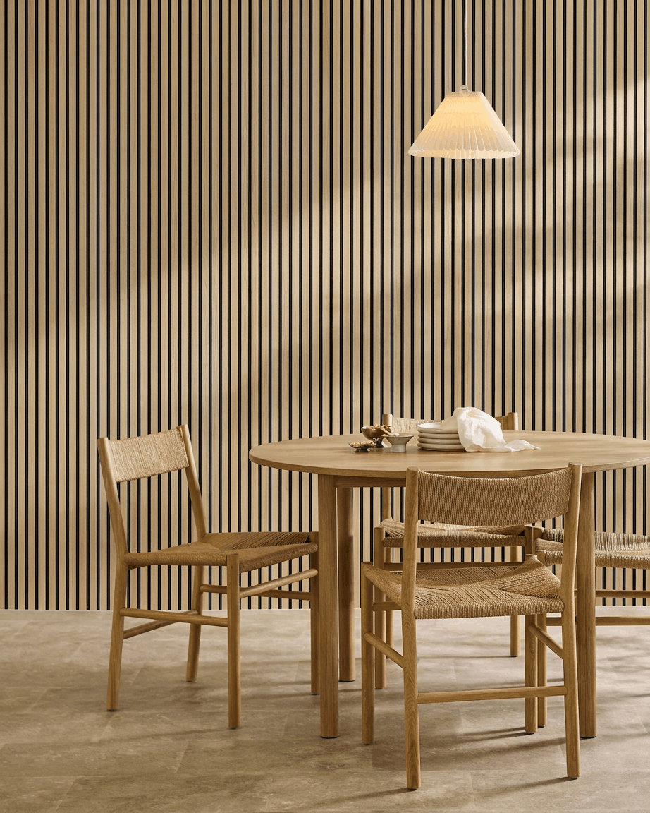 Comedor minimalista con una mesa redonda, sillas de mimbre, lámpara colgante y un WoodUpp Akupanel Classic Oak Panel acústico montado en la pared.