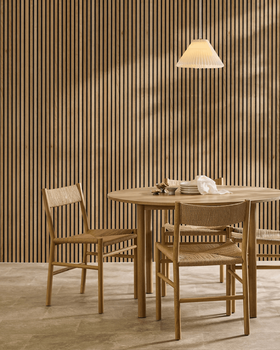 Comedor minimalista con una mesa de madera, dos sillas y un panel acústico de madera WoodUpp Akupanel en Rustic Brown Oak (300x60 cm), iluminado por una lámpara colgante.