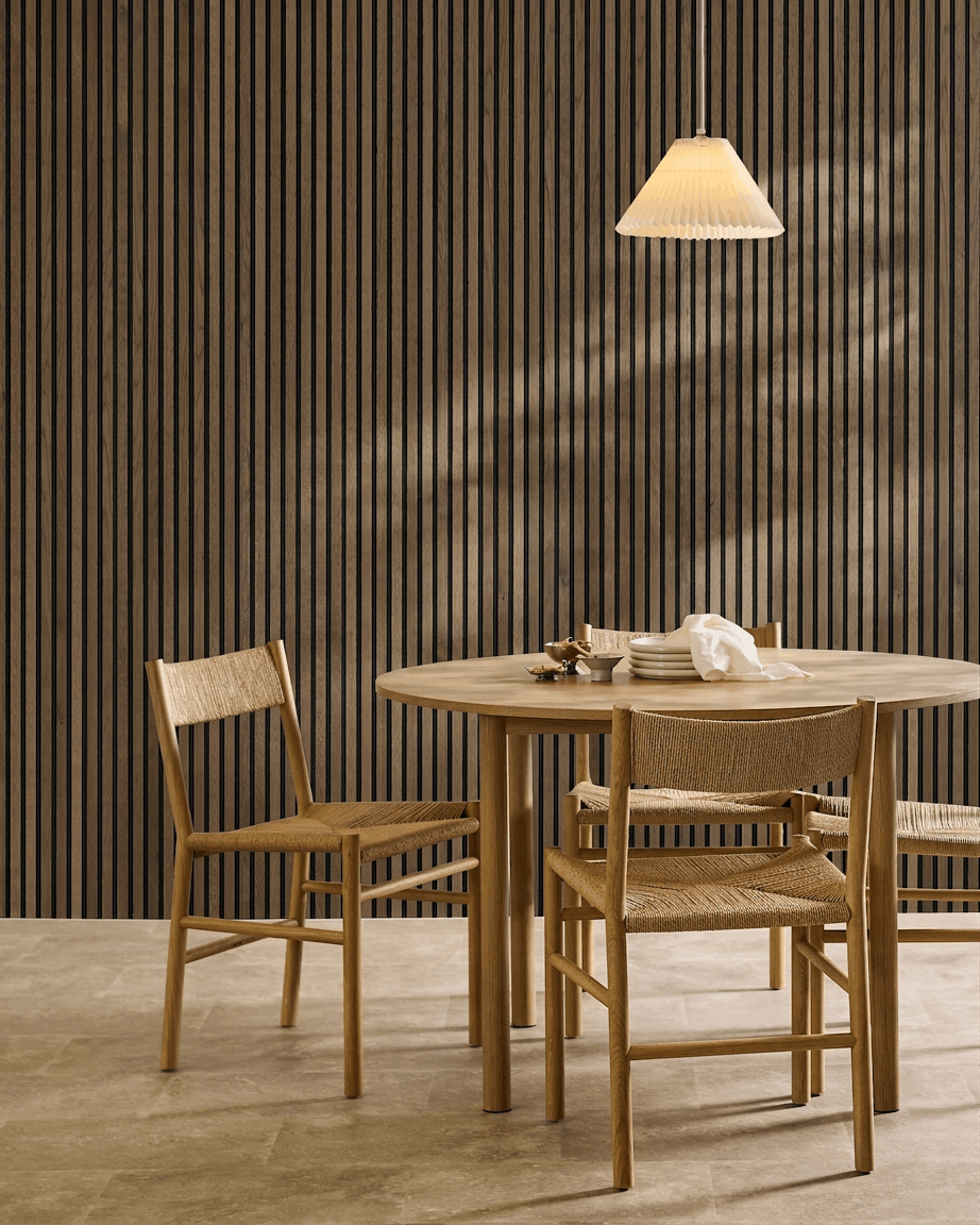 Comedor minimalista con mesa redonda, sillas tejidas y lámpara colgante, con un panel acústico de lamas WoodUpp Akupanel Rustic Grey Oak.
