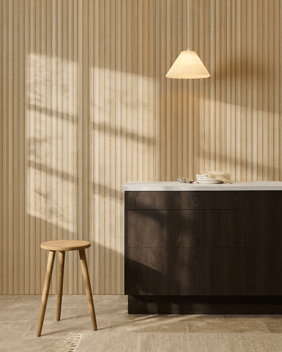 Cocina escandinava con el panel de listones acústicos WoodUpp Akupanel Classic Oak, armarios de madera oscura, taburete de madera, lámpara colgante y luz solar proyectando sombras.