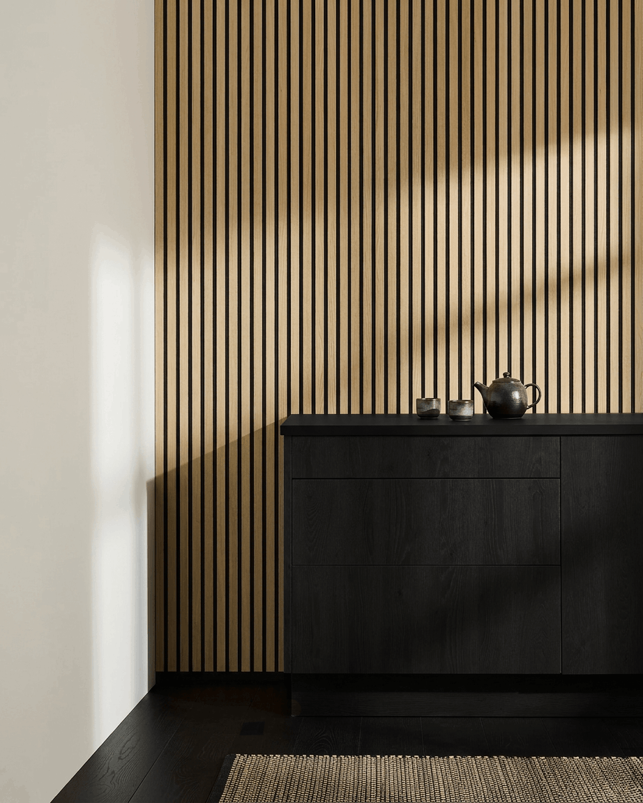 Cocina nórdica minimalista con WoodUpp Akupanel Panel acústico en roble clásico, armario black, tetera, tazas y alfombra tejida sobre suelo de madera oscura.