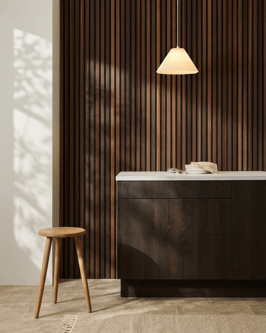 Interior minimalista con un taburete de madera, un armario de madera oscura, una lámpara colgante y un panel acústico de listones de madera WoodUpp Akupanel en Natural Wood/Smoked Larch.