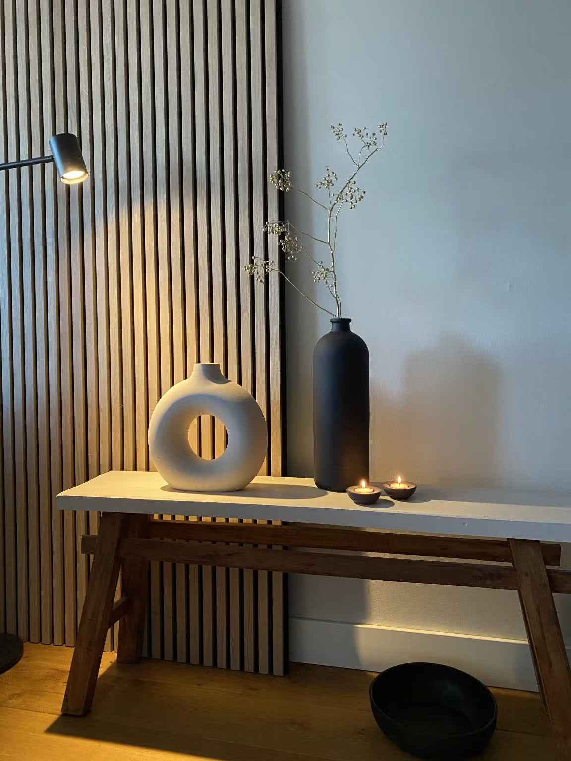 Decoración minimalista con un jarrón circular blanco, un jarrón alto negro con tallos secos, dos velas candelitas y un cuenco negro sobre un fondo de panel acústico de listones de madera de Akupanel.