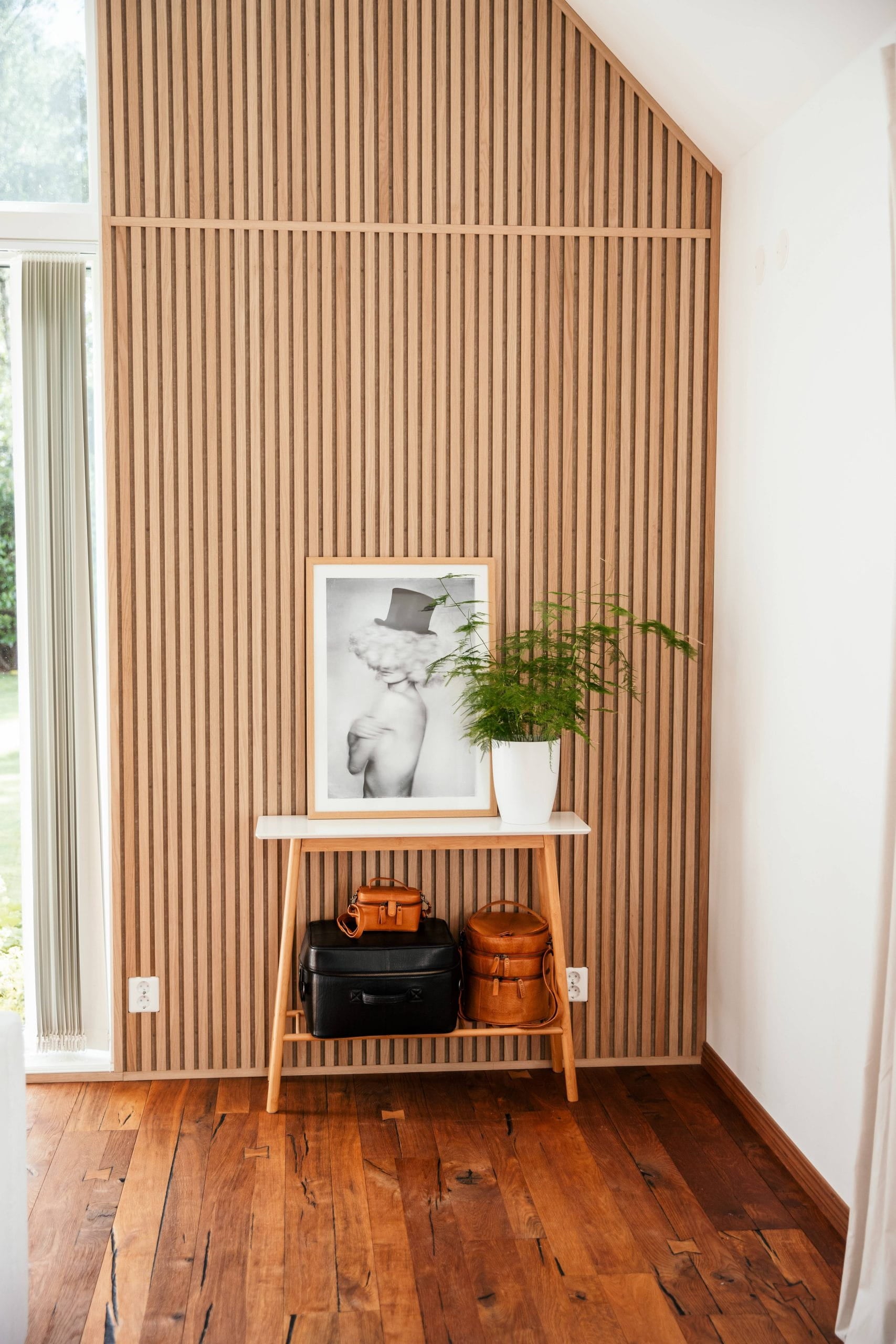 Una pequeña mesa de madera con bolsos, una foto enmarcada y una maceta se alza contra un panel de lamas acústicas nórdicas en una luminosa habitación con suelo de madera.