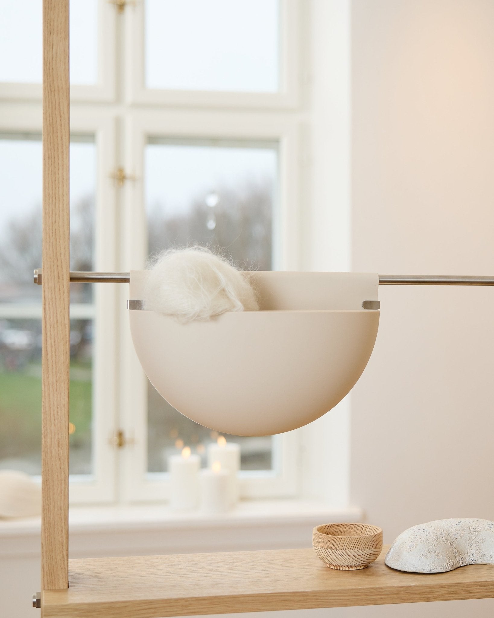Un cuenco blanco con un objeto mullido descansa sobre un estante de madera, junto a velas, decoración y un accesorio acústico WoodUpp Gro Soft Pocket BM2 en color claro beige, junto a la ventana.