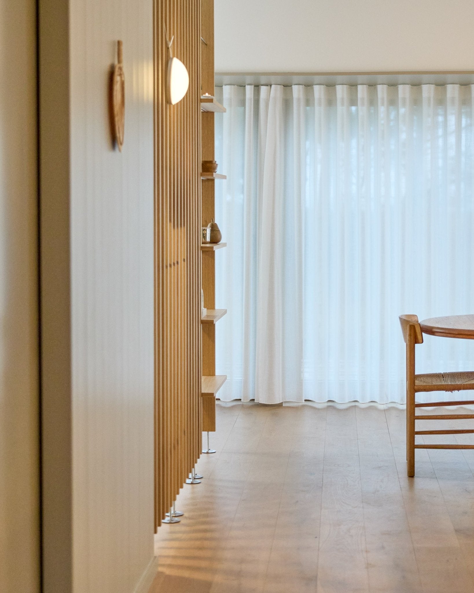Espacio minimalista con un separador de ambientes de suelo a techo WoodUpp Embrace, suelo de madera, estantes, una silla y cortinas translúcidas sobre ventanas large.