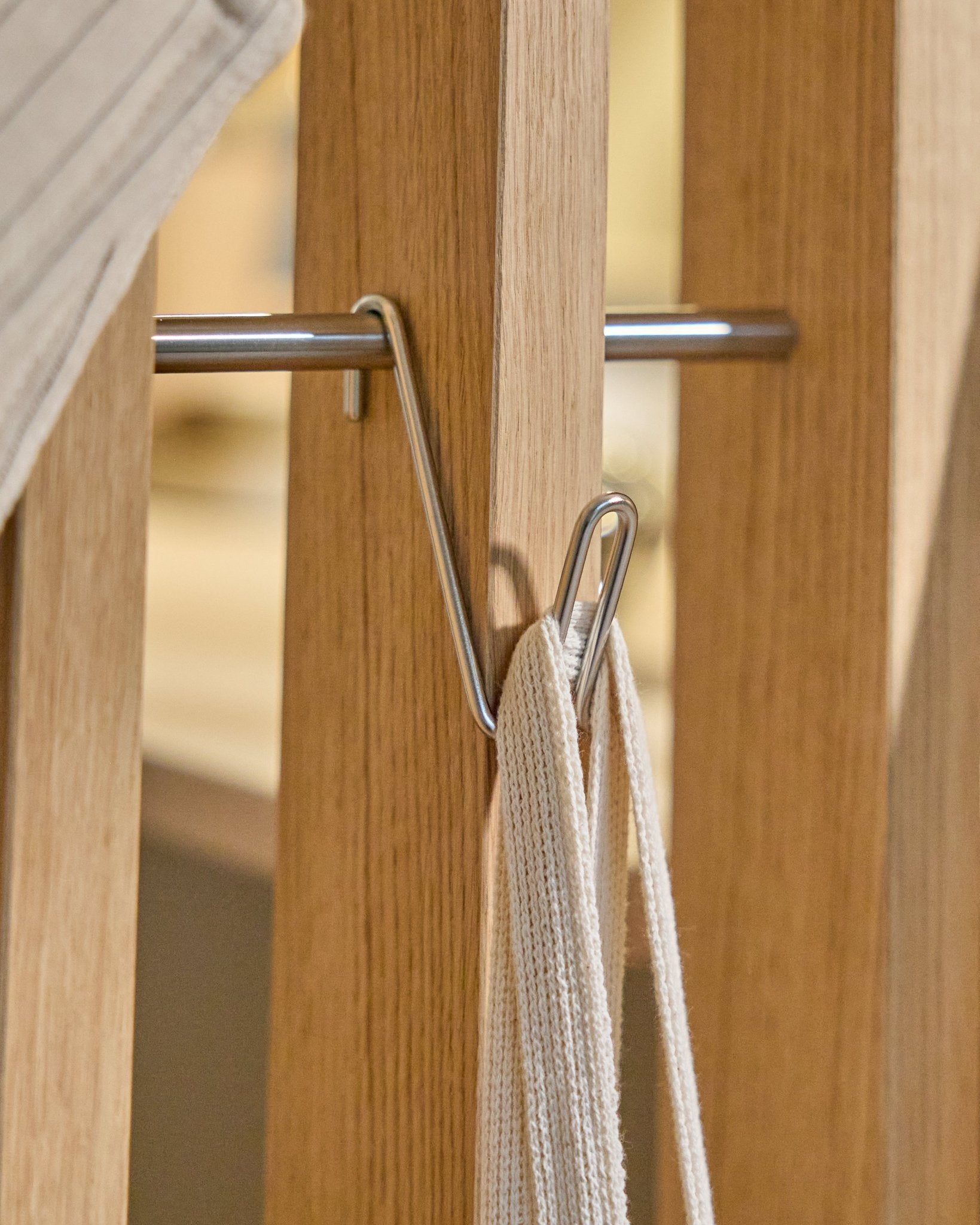 El Alva Wire Hook / BM1 (acero inoxidable Steel, corto) de WoodUpp está montado sobre un riel de madera Panel acústico y sostiene una correa blanca con varilla metálica; fondo beige.