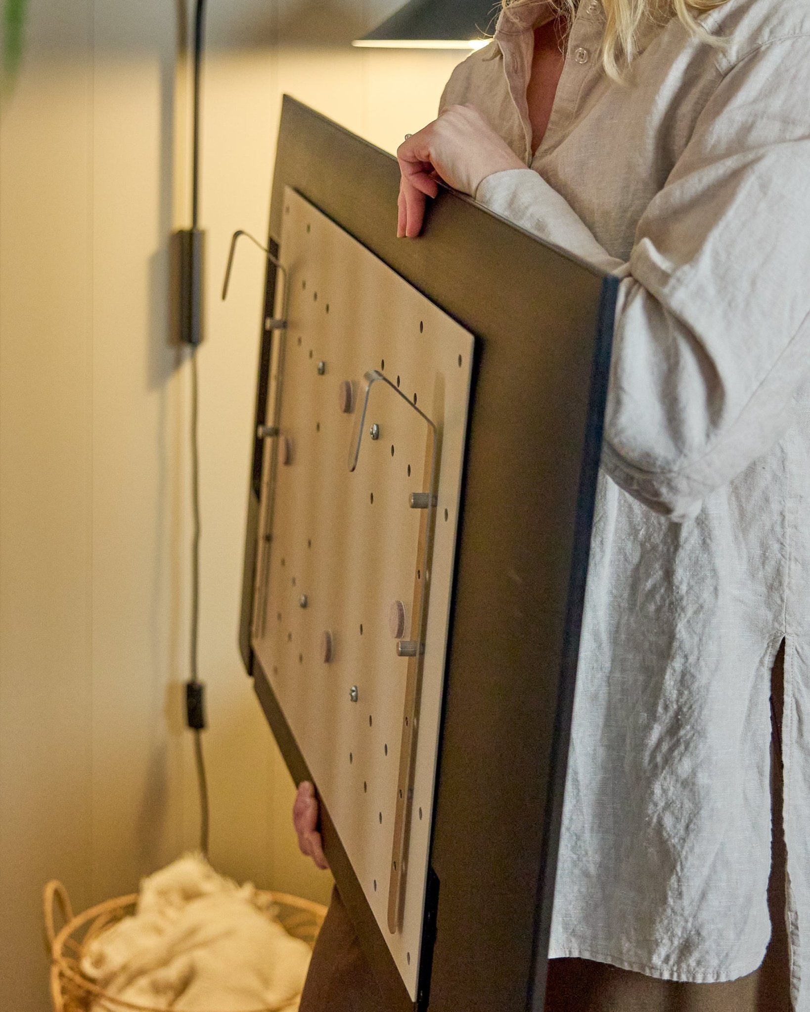 Una persona con camisa clara sostiene el WoodUpp Nell BM1 TV-Mount en beige, 64x44 cm, compatible con paneles acústicos nórdicos de madera, para instalaciones sin pared.