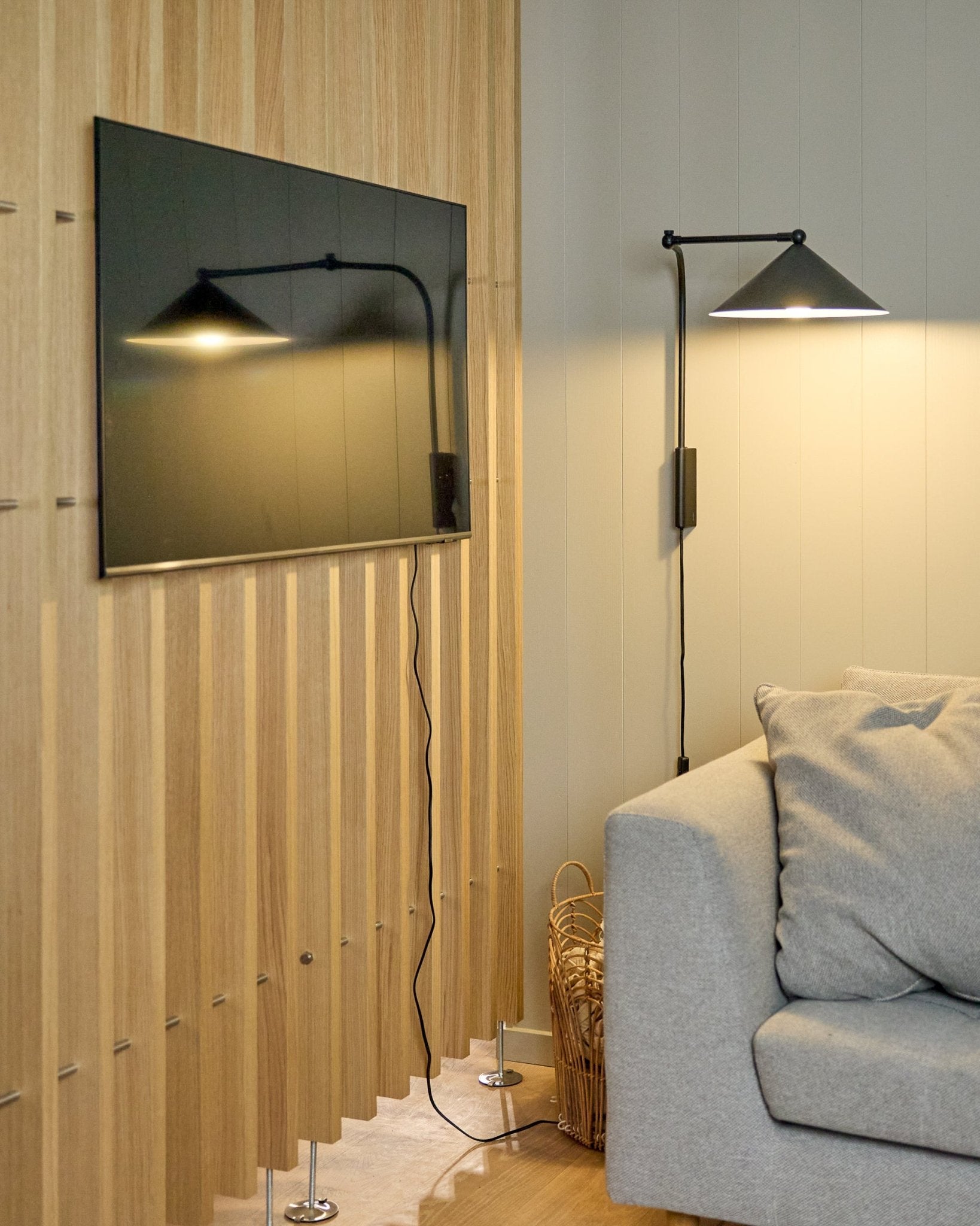 Un Nell | TV-Mount / BM1 de WoodUpp (Beige, 64x44 cm) sujeta un televisor de pantalla plana sobre un Panel acústico nórdico junto a un sofá gris; hay una cesta cerca.