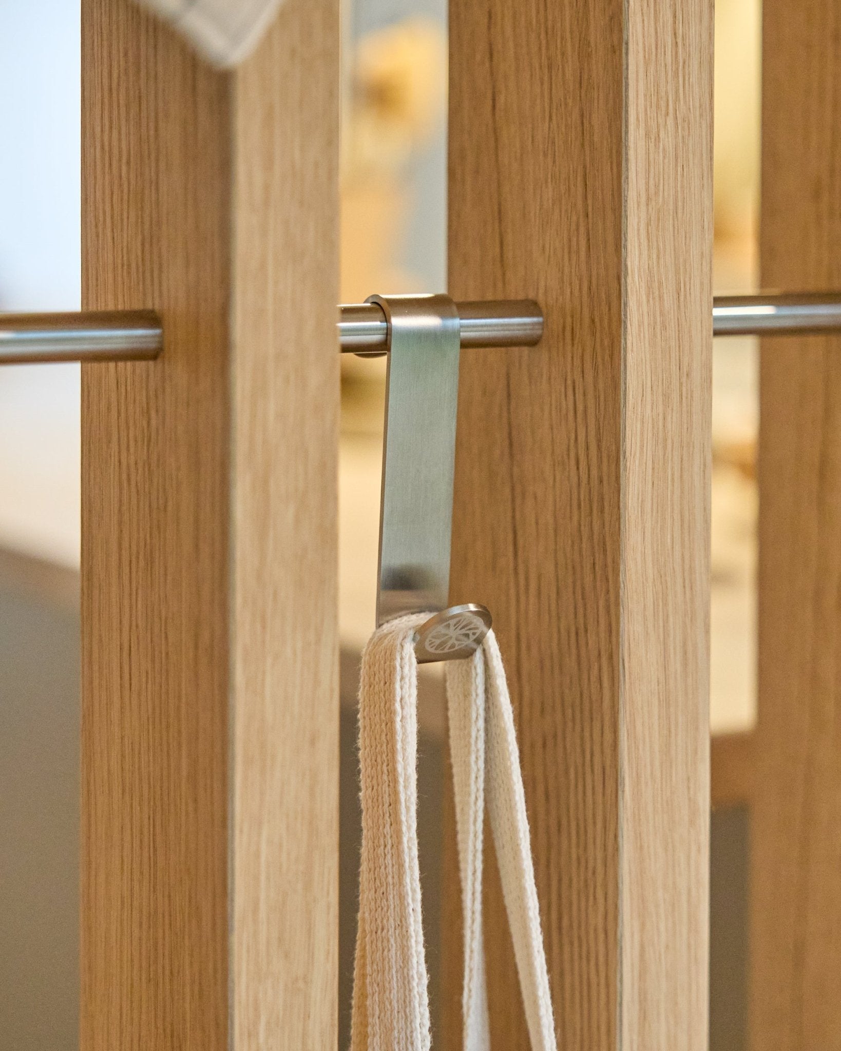 Un gancho en S Alva de WoodUpp en acero inoxidable steel sujeta una correa beige a una barra montada entre paneles acústicos de lamas de madera vertical. El juego incluye 2 ganchos.