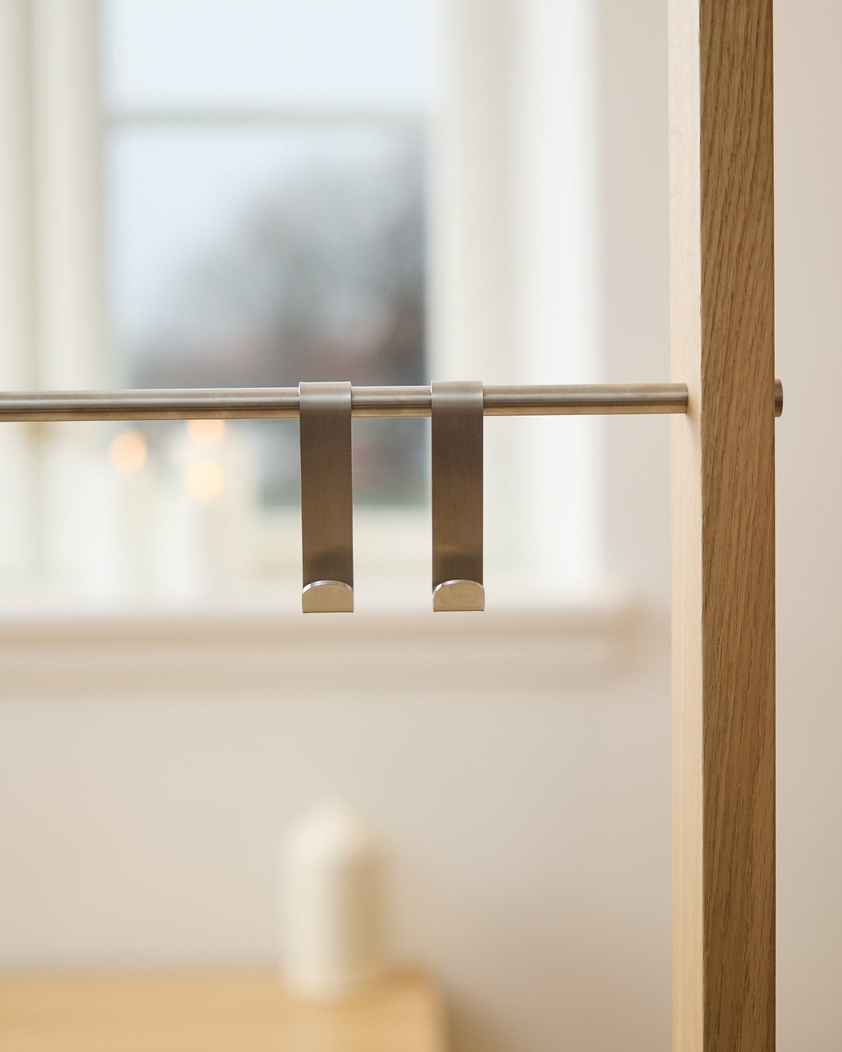 WoodUpp Alva S-Hook BM1  BM2 (2 uds.) en acero inoxidable steel, colgados de una barra para panel acústico de madera; al fondo, una ventana desenfocada y velas encendidas.